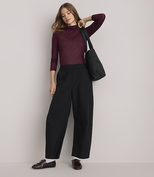 A woman in a burgundy turtleneck and black wide-leg pants stands against a plain white background, carrying a quilted black tote bag.