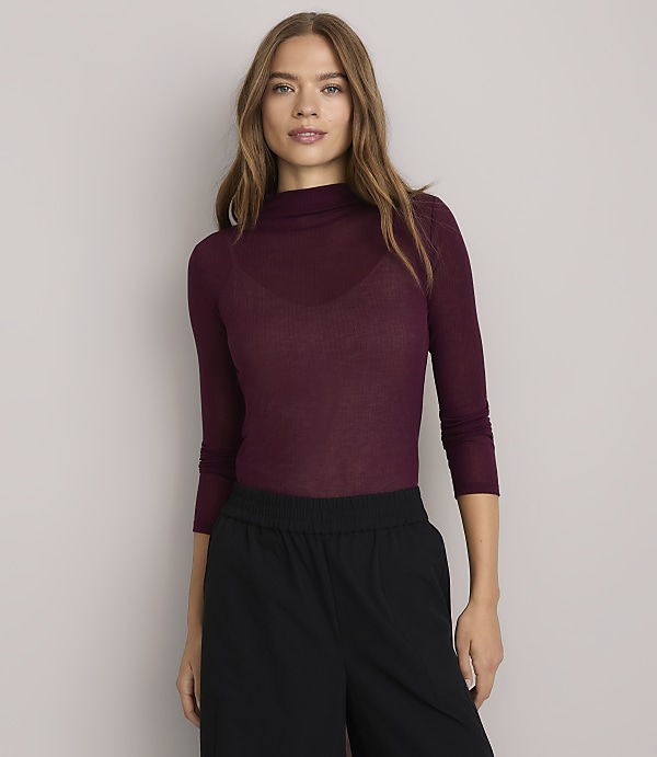 A woman in a burgundy turtleneck and black pants stands against a plain white background.