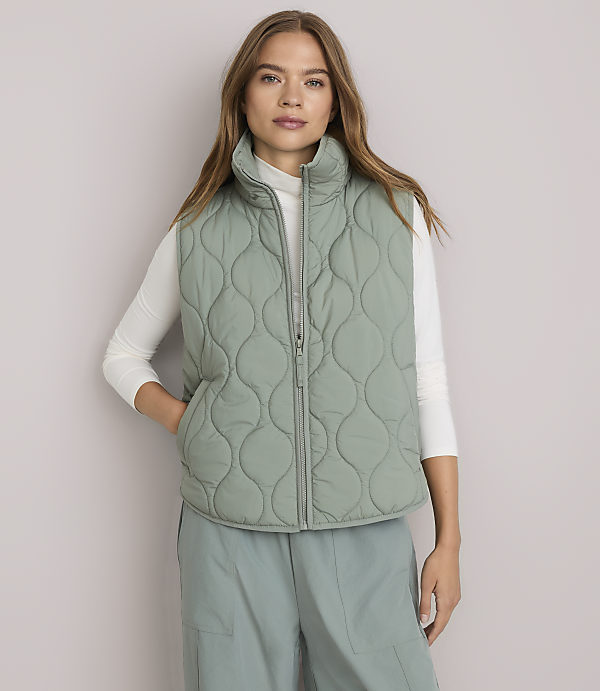 A woman in a light green quilted vest and white turtleneck stands against a plain white background.