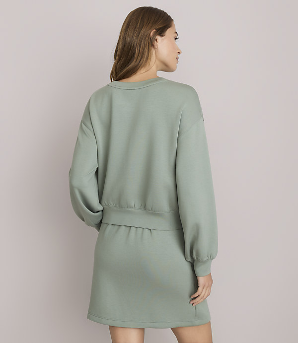 A woman in a sage green sweatshirt and skirt stands against a plain white background.