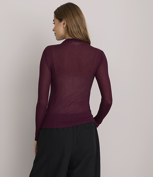 A woman in a sheer burgundy turtleneck and black pants stands against a plain white background.