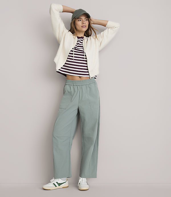 A young woman in a cream cardigan, striped t-shirt, and mint green pants stands with her hands behind her head against a plain white background.