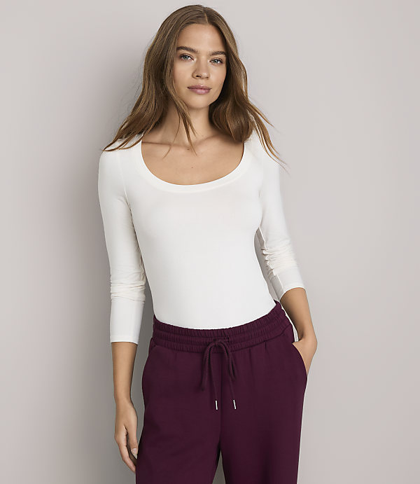 A woman in a white long-sleeved shirt and burgundy sweatpants stands against a plain gray background.