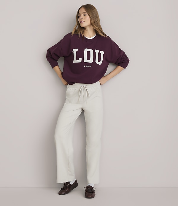 A young woman in a maroon sweatshirt and light-colored pants stands against a plain white background.