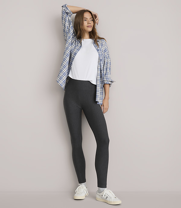 A woman in a white t-shirt, grey leggings, and a blue plaid shirt stands against a plain white background.