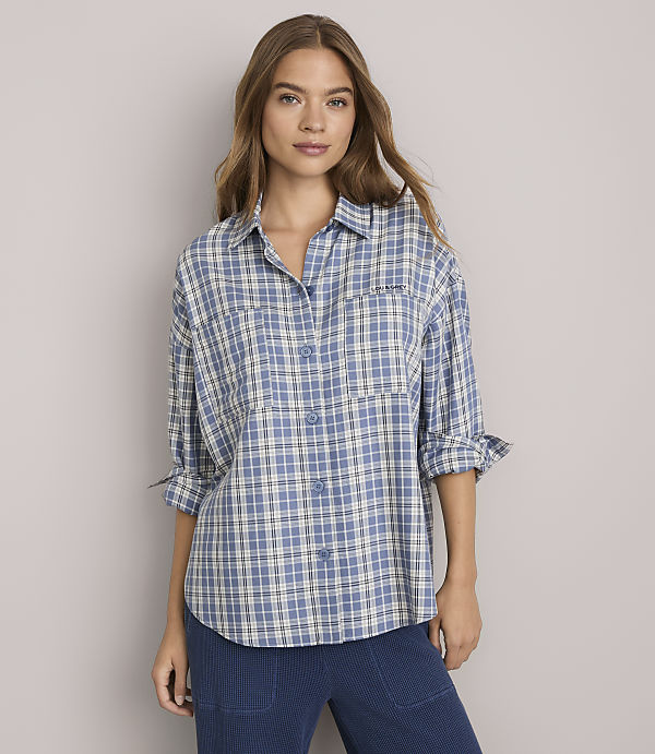 A woman in a blue plaid shirt stands against a plain gray background.