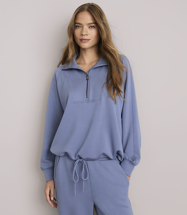 A woman in a blue quarter-zip sweatshirt and matching sweatpants stands against a plain gray background.