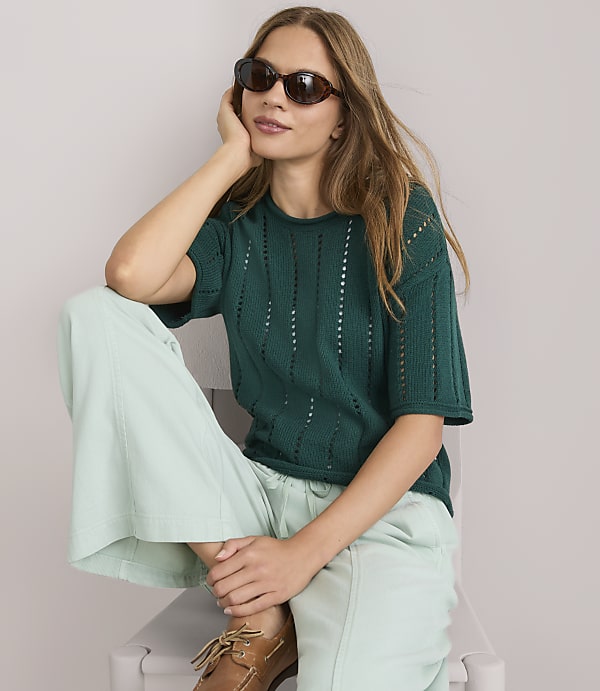 A young woman in a green v-neck sweater and light green pants stands against a plain gray background.
