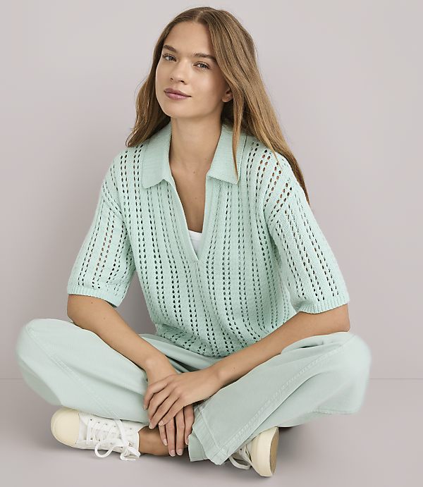 A young woman with long, light brown hair sits cross-legged in a light mint green collared knit shirt and matching wide-leg pants, against a plain white background.