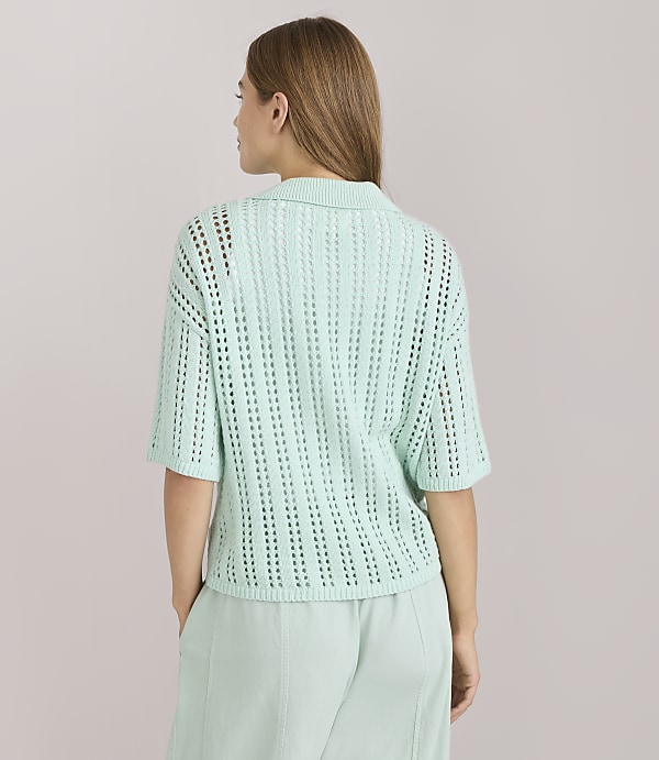 A woman in a light green collared knit shirt and matching pants stands against a plain white background.