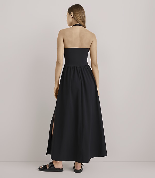 A woman in a black halter maxi dress with a slit stands against a plain white background.