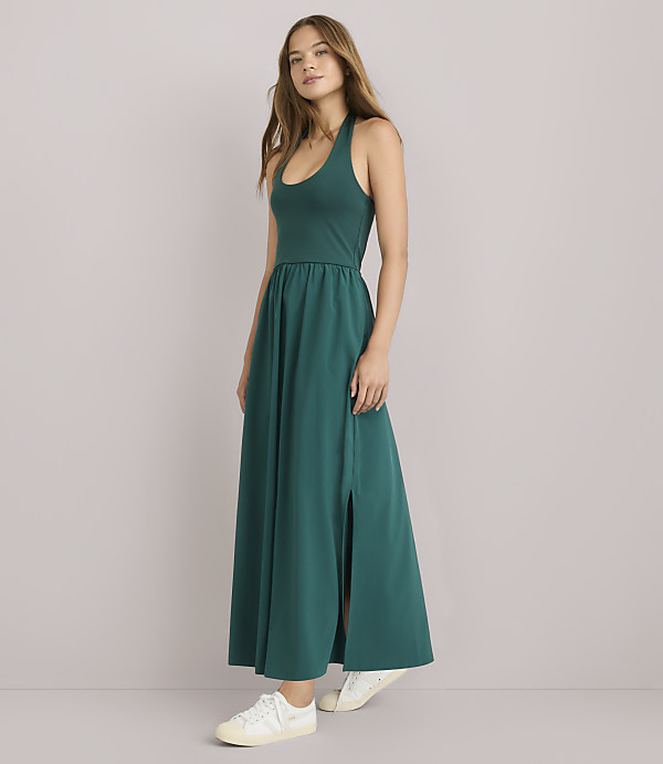 A woman in a teal maxi dress with a high slit and white sneakers stands against a plain gray background.