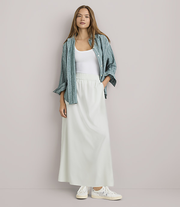 A woman in a white tank top, a green patterned shirt, and a long white skirt stands against a plain white background.