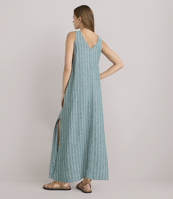 A woman in a teal striped maxi dress with side slits stands with her back to the camera against a plain white background.