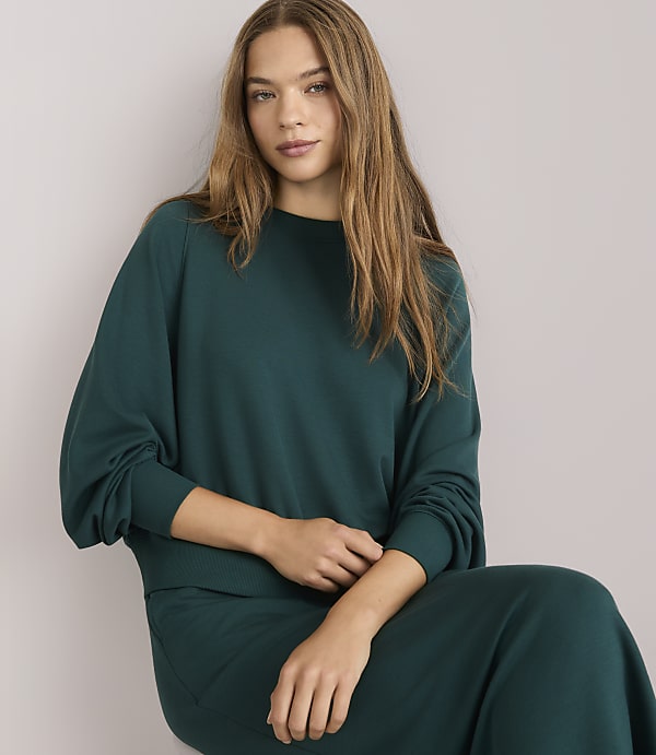 A woman in a teal sweatshirt and skirt stands against a plain white background.
