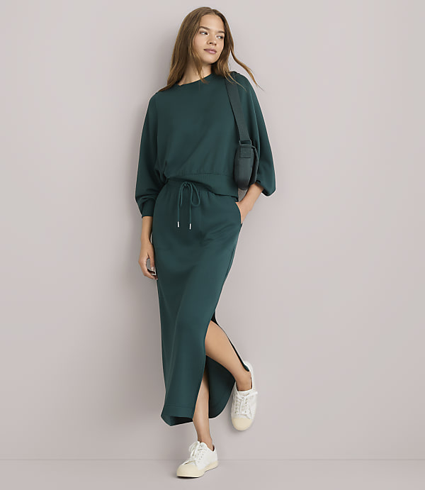 A woman in a teal sweatshirt and matching skirt with a side slit stands against a plain white background, wearing white sneakers and a small crossbody bag.