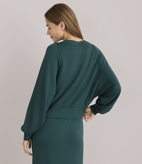 A woman in a teal sweatshirt with voluminous sleeves stands against a plain, light gray background.