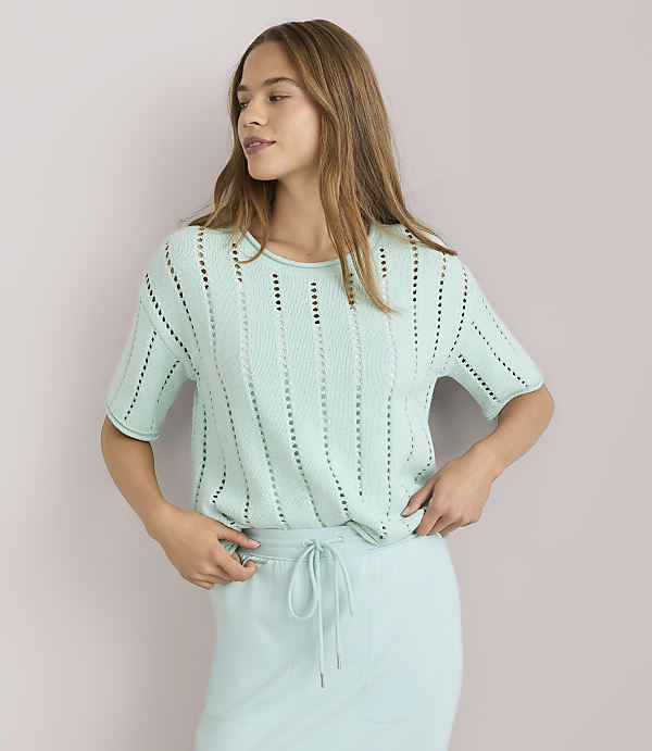 A young woman in a light green, short-sleeved, open-knit sweater and matching skirt stands against a plain white background.