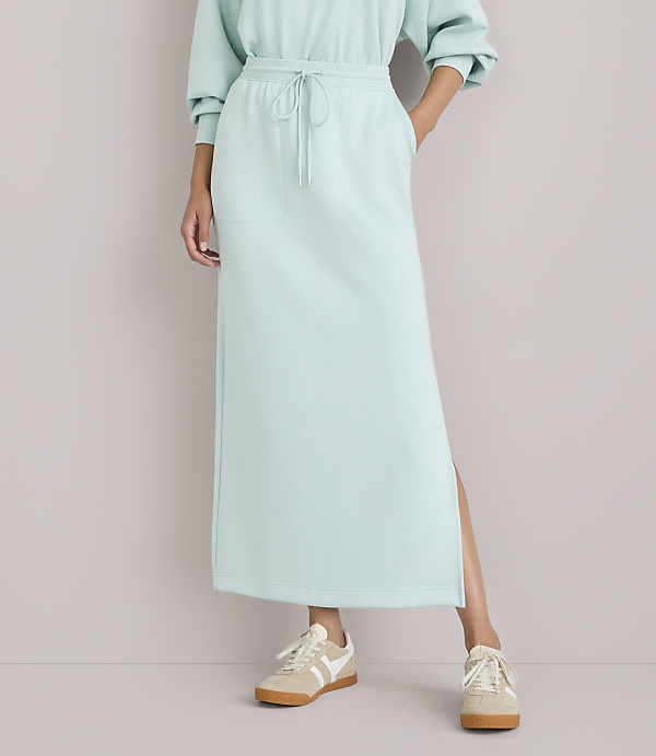 A person wearing a light blue sweatshirt and a matching maxi skirt with a slit, paired with beige sneakers, stands against a plain white background.
