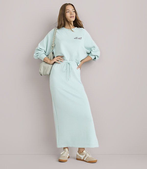 A young woman in a light blue sweatshirt and matching maxi skirt stands with her hands on her hips against a plain white background, a light gray purse slung over her shoulder.