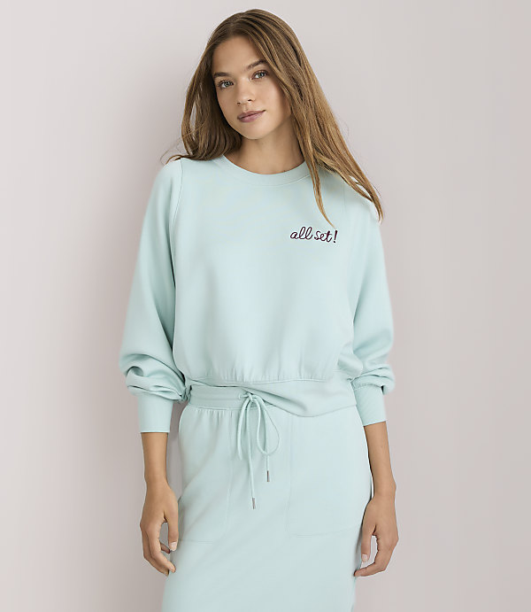 A young woman with light brown hair, wearing a light blue sweatshirt with "all set!" embroidered on it, poses with her arm behind her head against a plain white background.