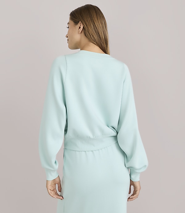 A woman in a light blue sweatshirt and skirt stands against a plain, light gray background.
