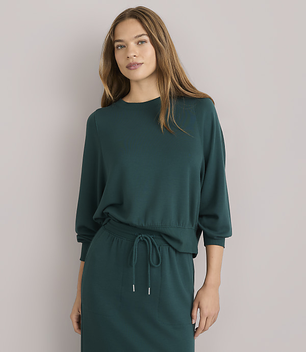 A young woman with long brown hair wears a dark green sweater and skirt, sitting against a plain white background.