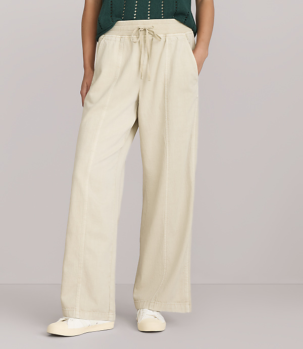 A person stands in the foreground wearing wide-leg cream-colored pants and a green sweater, against a plain white background.