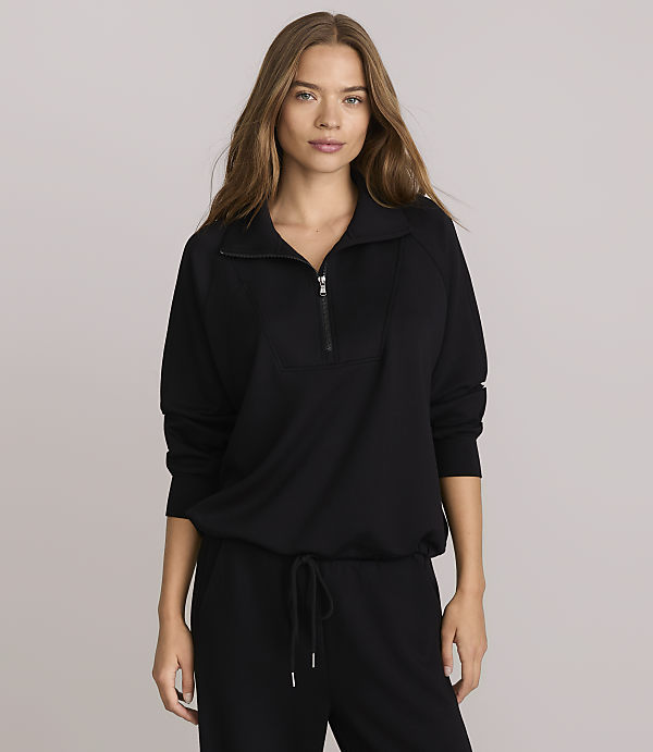 A woman in a black quarter-zip sweatshirt and sweatpants stands against a plain gray background.