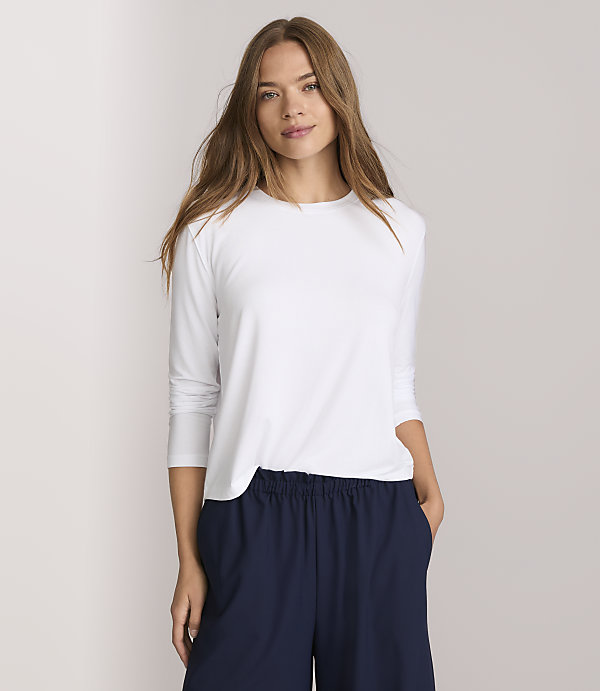 A woman in a white long-sleeved shirt and navy pants stands against a plain white background.