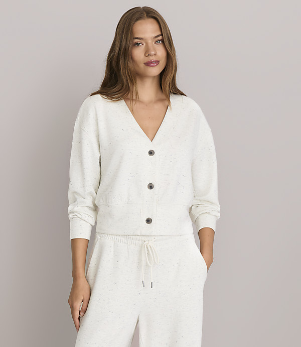 A woman in a cream-colored cardigan and matching pants stands against a plain gray background.