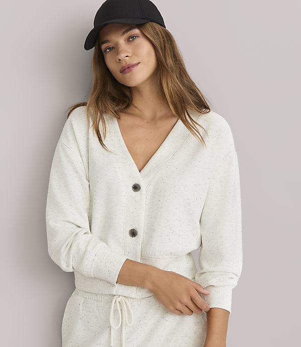 A young woman in a light-colored cardigan and sweatpants, wearing a black baseball cap, poses against a plain white background.