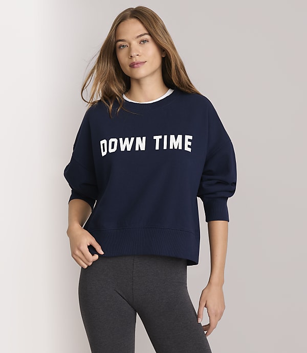 A young woman in a navy sweatshirt with "DOWN TIME" printed on it and dark leggings stands against a plain white background.