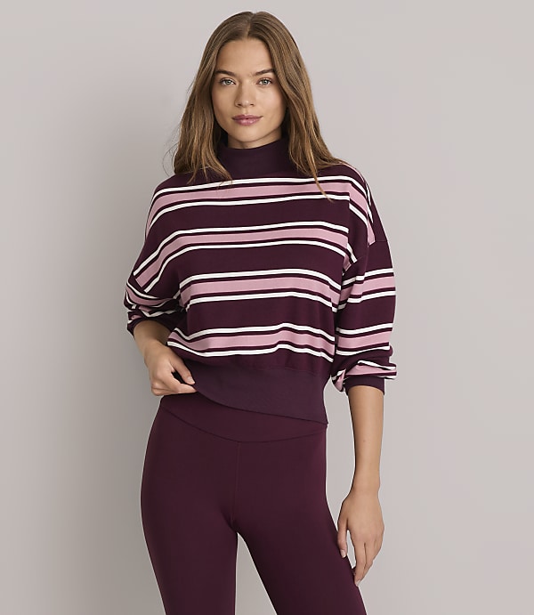 A woman in a striped mock-neck sweatshirt and burgundy leggings stands against a plain gray background.