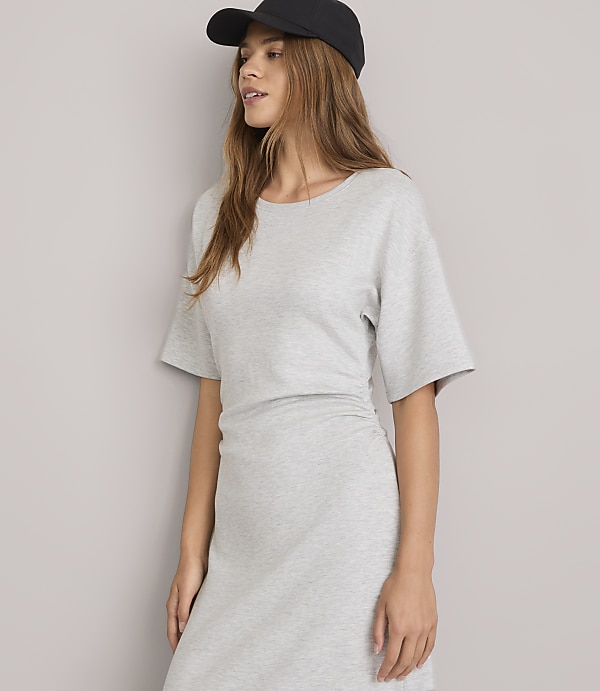 A young woman in a grey dress and black baseball cap stands against a plain grey background.