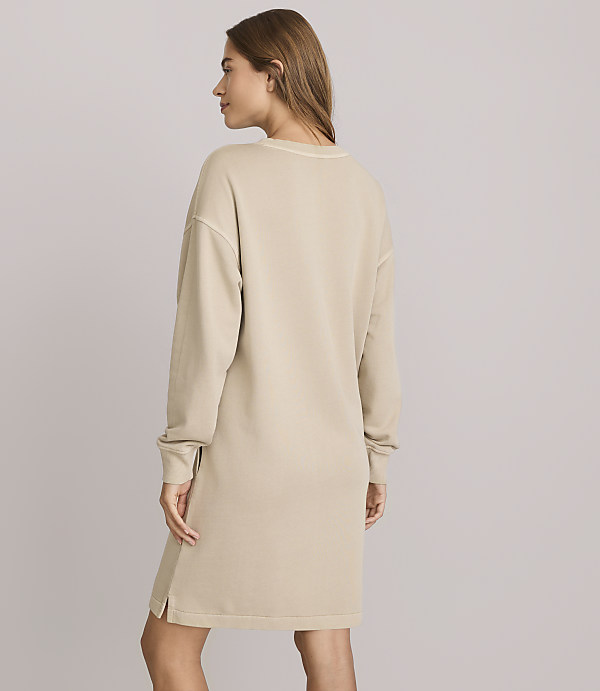 A woman in a beige sweatshirt dress stands against a plain, light gray background.