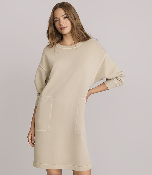 A woman in a beige sweatshirt dress with pockets stands against a plain gray background.