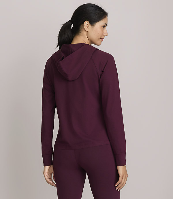 A woman in a burgundy hoodie and leggings stands facing away from the camera against a plain gray background.