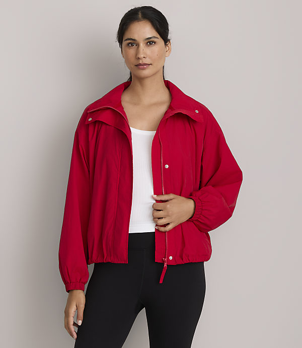 A woman in a red jacket and white tank top stands against a plain gray background.