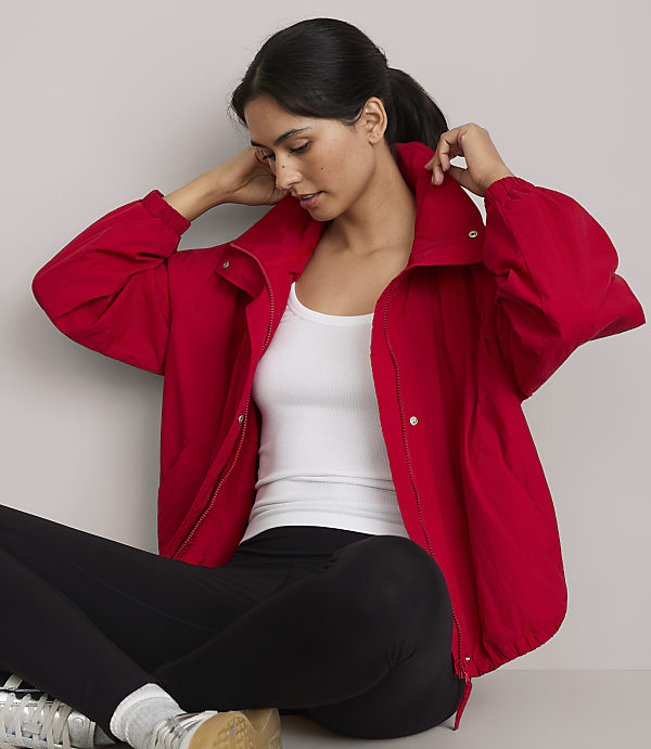 A woman in a red jacket and black leggings sits against a plain gray background.