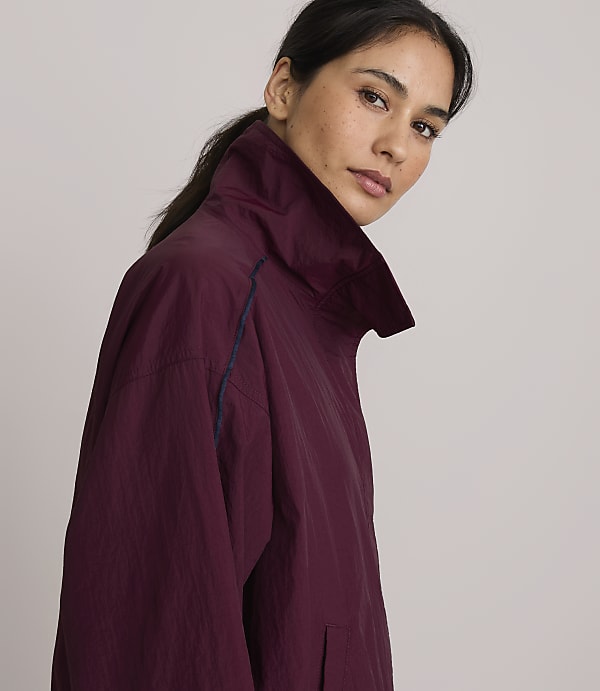 A woman in a burgundy jacket with a high collar is shown in profile against a plain, light gray background.