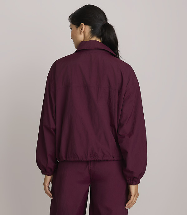 A woman in a maroon jacket and pants stands against a plain, light purple background.