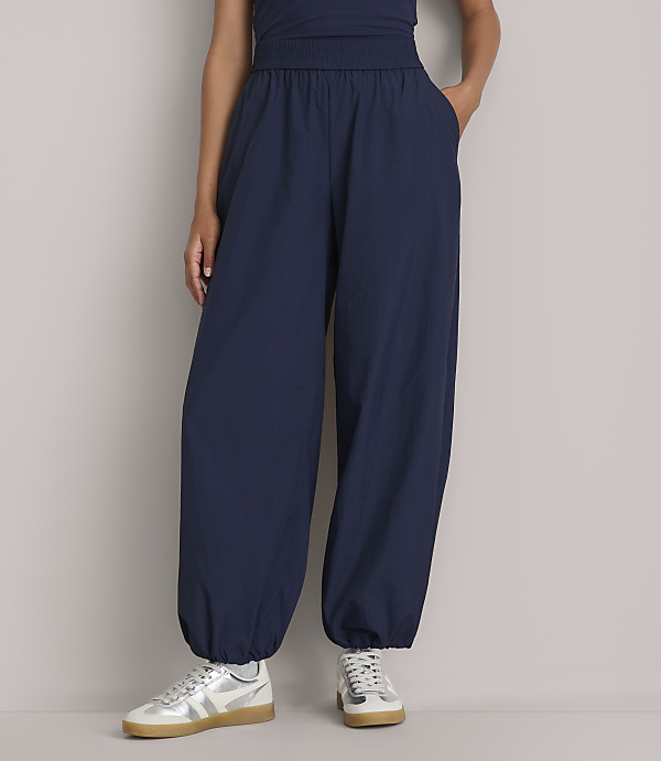 A person stands in dark blue, baggy pants with cinched ankles and white sneakers against a plain gray background.