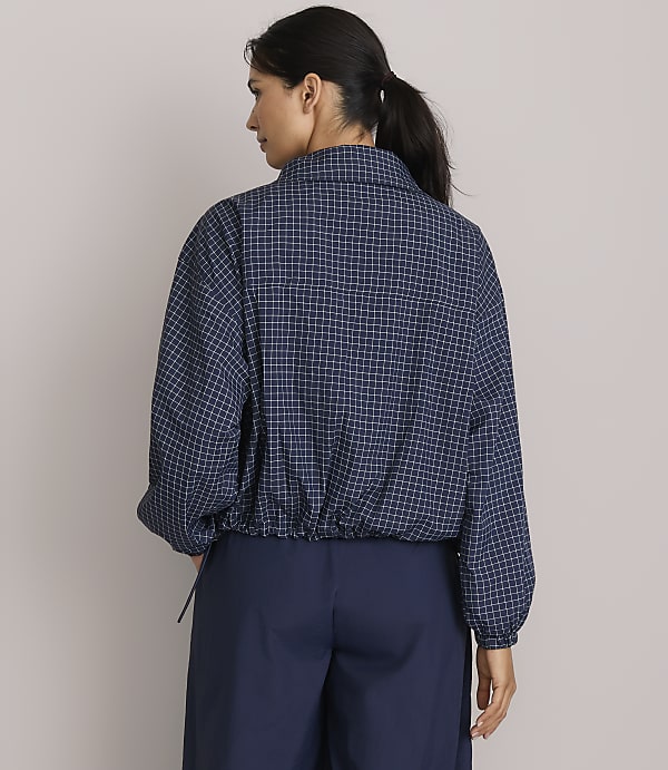 A woman in a checkered jacket and navy pants stands against a plain gray background.