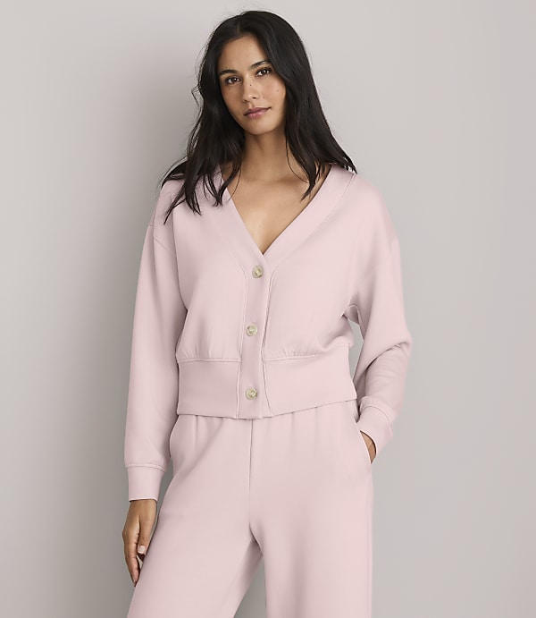 A woman in a pink cardigan and matching pants stands against a plain gray background.