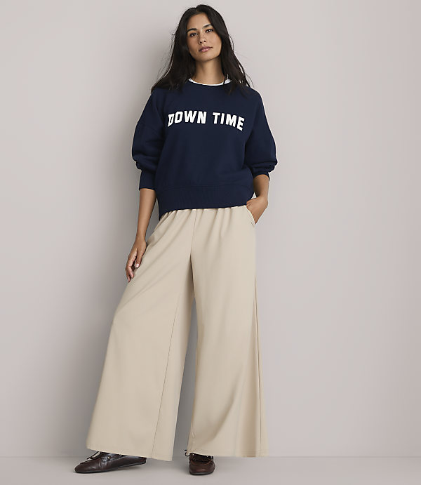 A woman in a navy sweatshirt and wide-leg pants stands against a plain gray background.