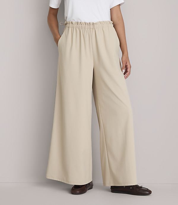 A person stands in wide-leg beige pants and brown shoes against a plain gray background.