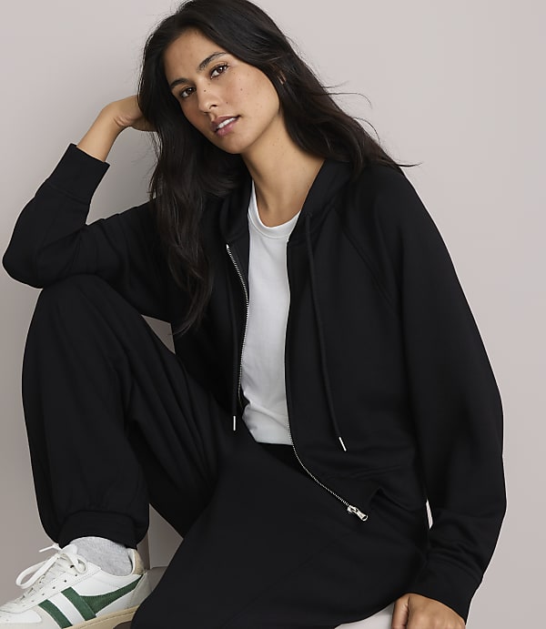 A woman in a black zip-up hoodie and sweatpants poses against a plain, light-colored background.