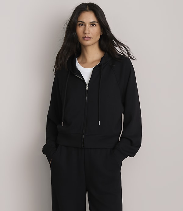 A woman in a black zip-up hoodie and sweatpants stands against a plain, light gray background.