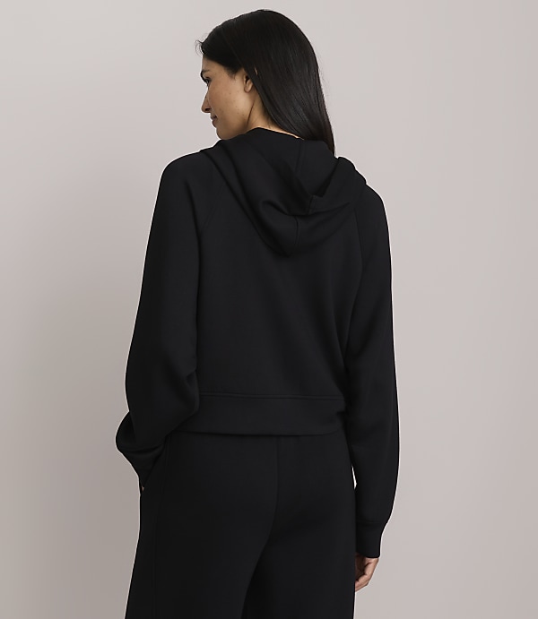 A woman in a black hoodie and pants stands with her back to the camera against a plain gray background.